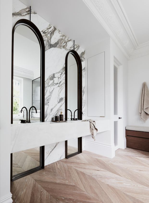 Bright, refined bathroom interior featuring a long white marble double vanity with black arched-framed mirrors and black gooseneck faucets. The room has a dramatic veined marble feature wall, pale herringbone timber flooring, white painted panelled walls, ornate decorative ceiling moulding, and a dark timber vanity cabinet.
