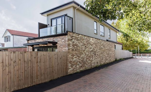 Exterior view of a contemporary two-storey residential building, showing a pale rendered upper level, extensive variegated brickwork at ground level, dark metal-framed windows and balcony balustrade, and a low-pitched roof. A timber privacy fence and brick-paved driveway or shared access area occupy the foreground, with neighbouring homes and mature trees around the property.
