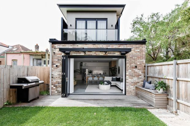 Rear exterior view of a contemporary two-storey home with a brick lower-level extension, wide open bi-fold glazing, and an upper balcony with frameless glass balustrade. The ground-floor opening reveals a bright living and dining area with a kitchen beyond. A timber deck, built-in planter seating, barbecue, fenced yard, and lawn complete the backyard setting.