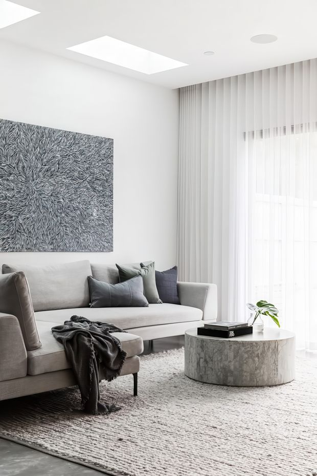 A bright, refined living room with a large pale sectional sofa, dark and neutral cushions, a textured grey rug, and a round cylindrical marble coffee table. Full-height sheer curtains cover the window wall, while recessed ceiling lights and a large rectangular skylight provide abundant daylight. A monochrome abstract artwork adds visual interest to the otherwise soft, neutral scheme.
