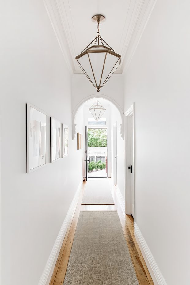 Bright, narrow residential hallway with white walls, decorative ceiling cornices, timber floorboards, and a long neutral runner rug. Framed artwork lines the left wall, while white doors open off the right side. A large bronze-toned geometric pendant light hangs centrally, and an arched opening leads to a glazed exterior door with greenery visible beyond.