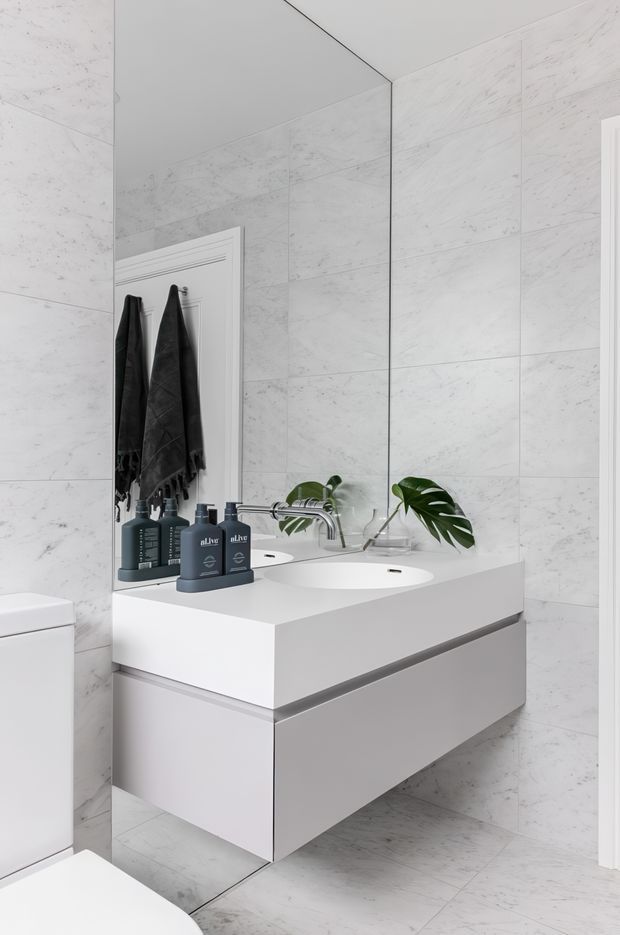 Bright contemporary bathroom featuring large white marble-look wall and floor tiles, a floating double-drawer white vanity with integrated twin basins, wall-mounted chrome tapware, and a large frameless mirror. Dark towels and green foliage provide restrained contrast.