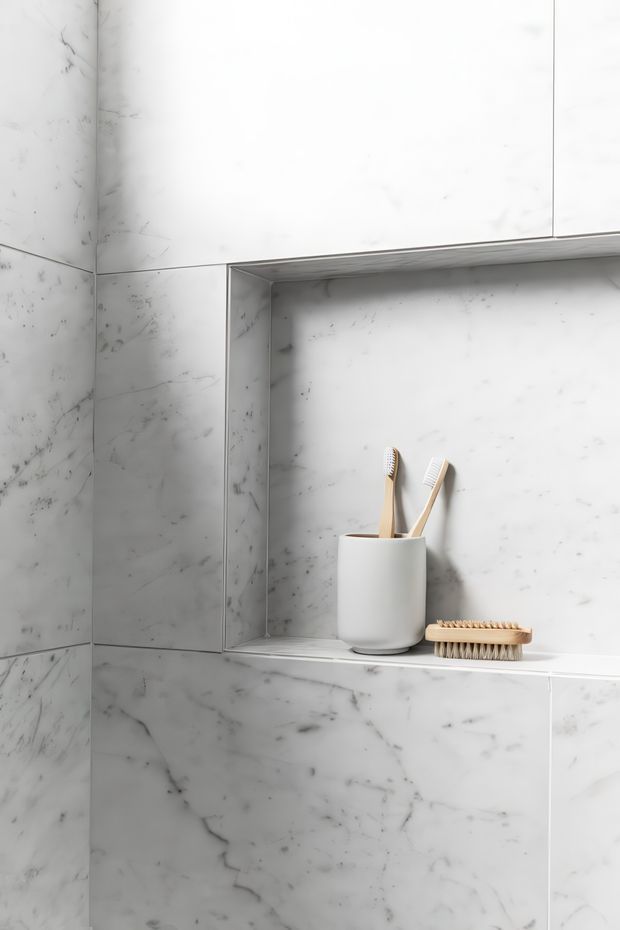 Close-up of a minimalist bathroom shower or wet-area wall clad in white marble-look tiles, featuring a recessed wall niche with a white cylindrical cup holding toothbrushes and a wooden bath brush. The space is bright, clean, and monochromatic with subtle grey veining.