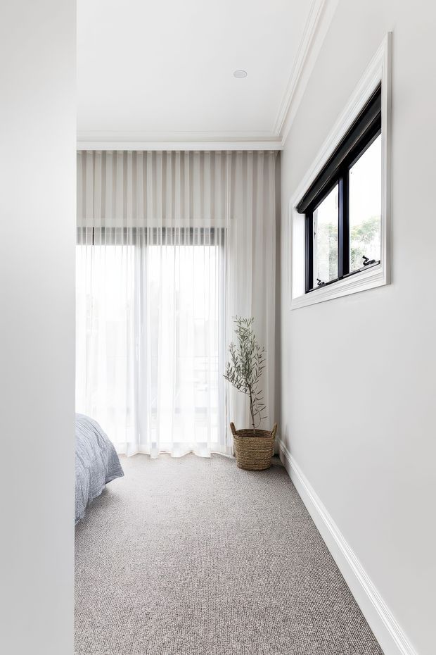 Bright, narrow bedroom corner with pale grey carpet, white walls and ceiling, decorative ceiling cornice, sheer floor-to-ceiling curtains, and a black-framed opening window with white trim. The edge of a bed is visible at left, while a woven basket planter with a slender leafy plant sits in the corner. The room has a clean, understated contemporary appearance with abundant natural light.