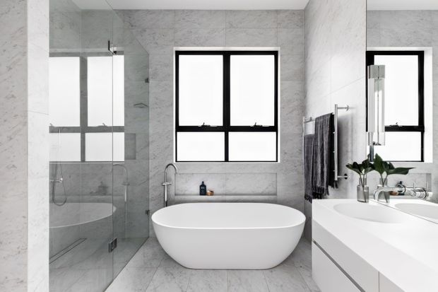 Bright contemporary bathroom clad in light grey marble-look surfaces, with a freestanding white oval bathtub, glass-enclosed shower, wall-mounted towel radiator, black-framed windows, and a white double vanity with integrated basins. The clean monochromatic scheme is accented by dark window frames and minimal greenery.