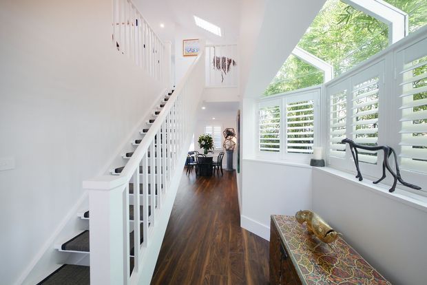 Bright, airy interior view of a residential stairway and landing with white painted balustrades, dark timber flooring, and extensive white plantation shutters. The staircase rises to an upper level beside a tall windowed area, while a small dining or sitting area is visible at the far end. A patterned console table and decorative objects occupy the foreground.
