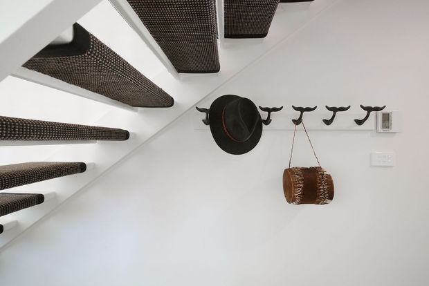 A compact white entry or hallway wall beneath a staircase. The stair treads have dark woven or textured undersides, while the wall features a row of black sculptural hooks holding a dark hat and a small woven hanging basket. White electrical controls are visible on the right.