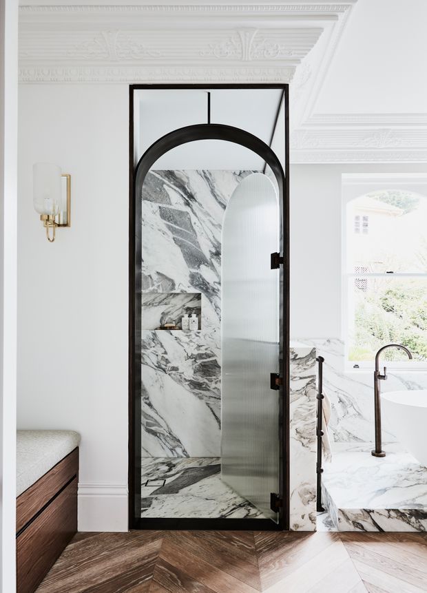 A refined traditional bathroom featuring an ornate white decorative ceiling, white walls, and a dramatic marble-clad shower enclosure with a black metal-framed arched ribbed-glass door. A freestanding white bathtub with dark metal tapware sits beside the shower, while a timber vanity and chevron timber flooring add warmth. The space combines heritage detailing with contemporary fixtures and bold natural stone.