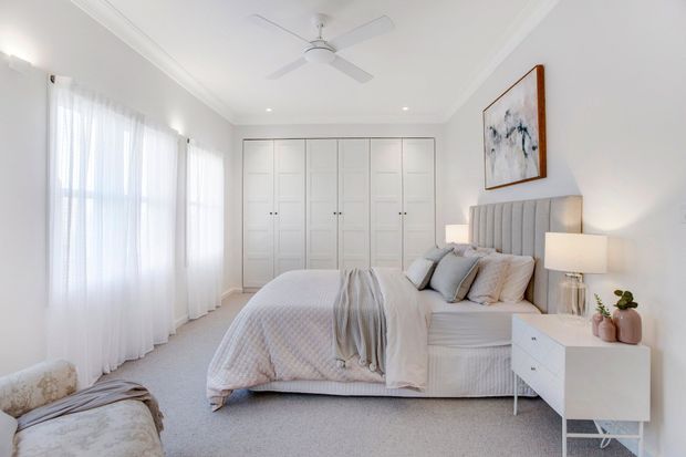 Bright, serene bedroom with a soft neutral palette, white built-in wardrobes, floor-to-ceiling sheer curtains, a large upholstered bed with layered bedding, matching bedside tables and lamps, and a ceiling fan. The room has carpeted flooring, simple cornice detailing, and restrained contemporary furnishings.