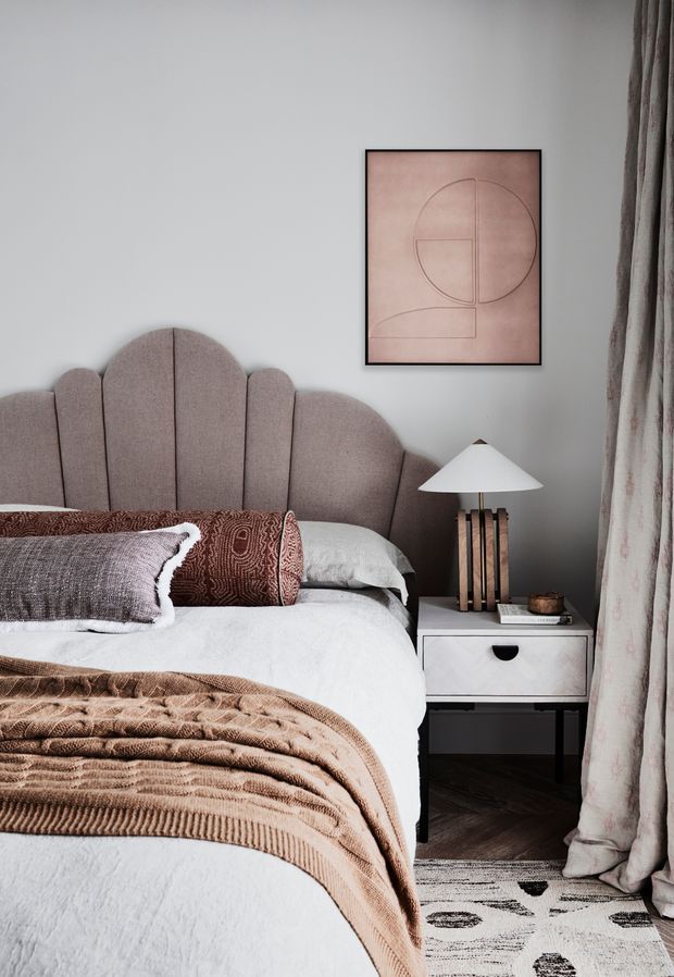 A warm, softly styled bedroom with a scalloped upholstered headboard, white bedding, layered taupe and brown textured throws and cushions, and a compact white bedside table with a sculptural timber-based lamp. A muted geometric artwork hangs above the table, while full-length patterned curtains frame the right side. Dark timber flooring and a partially visible patterned rug complete the room.