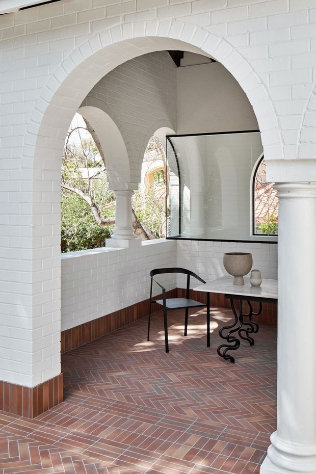 A bright, enclosed architectural terrace or verandah with white-painted brick walls, large rounded brick arches, classical columns, and curved glazed screens. The floor is finished in a red-brown herringbone tile pattern with a darker tiled perimeter skirting. A small stone-topped table, black sculptural metal table base, black chair, and ceramic vessels create a refined seating nook. Mature greenery is visible beyond the arches.