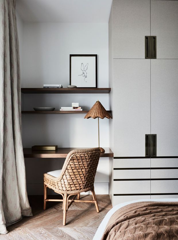 A calm, refined bedroom corner featuring a built-in home office nook with dark timber open shelving, pale cabinetry, a woven rattan armchair, a slim floor lamp with a natural woven shade, framed line art, and light timber flooring. The edge of the bed and textured neutral bedding are visible in the foreground, while full-height curtains frame the left side.