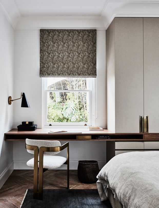 A refined bedroom workspace with a compact built-in timber desk running beneath a window, paired with a sculptural upholstered chair. Floor-to-ceiling built-in cabinetry occupies the right side, while a partially visible bed sits in the foreground. The room features white walls and ornate ceiling cornices, dark timber parquet flooring, a patterned Roman blind, brass wall lighting and hardware, and a restrained contemporary palette.