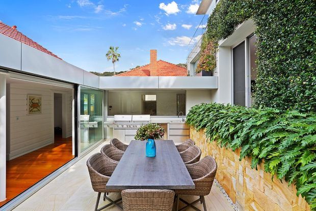 A bright contemporary courtyard dining area with a large outdoor timber dining table surrounded by woven chairs. The space connects to a compact outdoor kitchen or barbecue zone beneath a flat-roofed canopy, with glazed doors opening to an interior room. A textured stone retaining wall supports lush fern and hedge planting, while an adjacent dwelling has a red tiled pitched roof visible beyond the courtyard.
