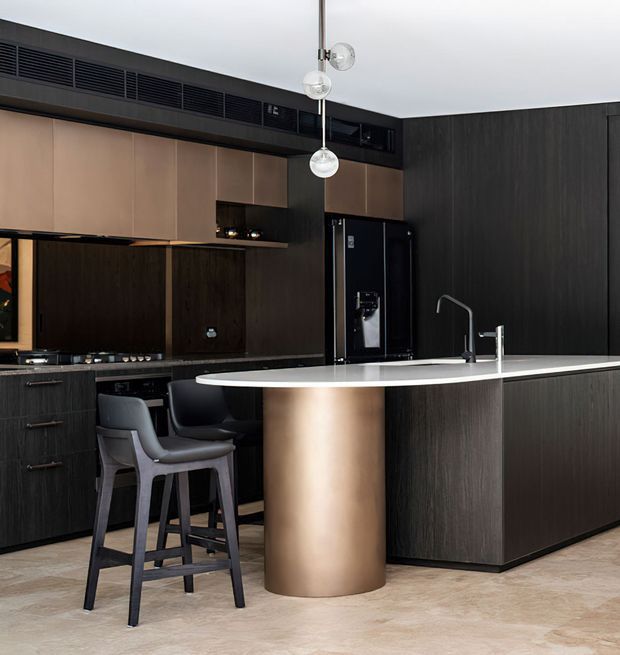 A sophisticated contemporary kitchen with a large dark timber island, pale stone benchtop, rounded bronze-toned cylindrical feature end, integrated dark cabinetry, black appliances, and two sculptural pendant lights. Two bar stools sit at the island, with a sink and minimalist tapware visible. The palette is dark, warm, and refined.
