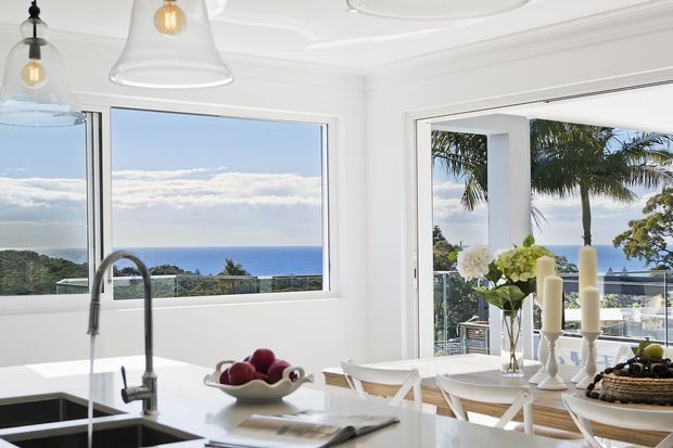 Bright coastal kitchen and dining area with white cabinetry and surfaces, a large island sink, pendant lights, and expansive windows and glazed doors overlooking a blue ocean horizon and palm-lined landscape. A timber dining table with white cross-back chairs sits beside the kitchen.