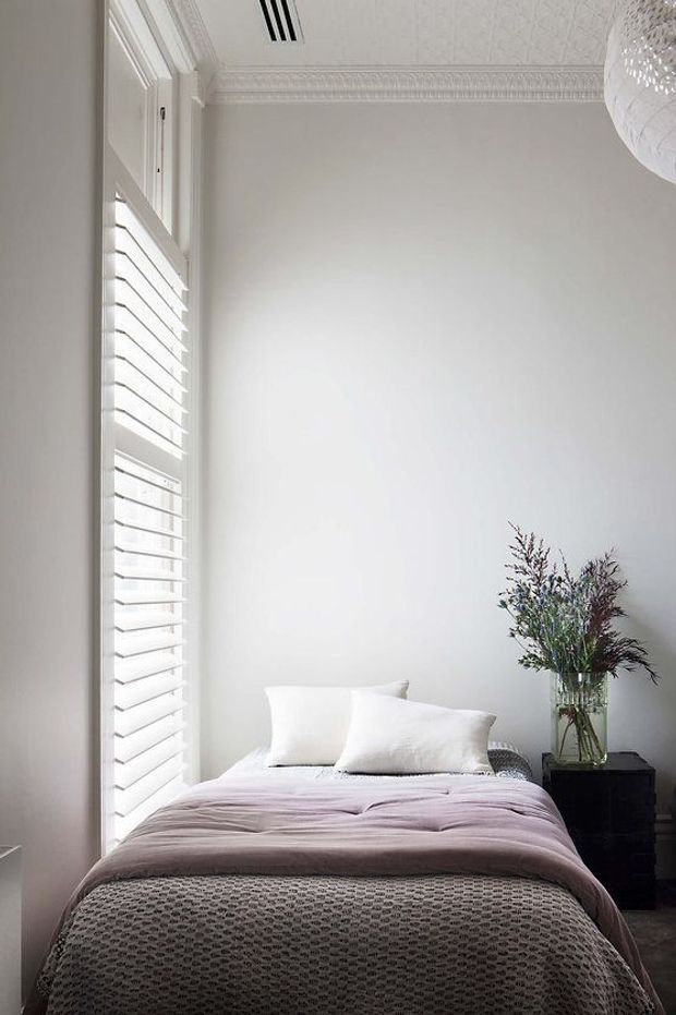 A bright, compact bedroom featuring a neatly made bed with soft white pillows, a muted mauve-gray throw, and a textured gray bedspread. Tall white plantation shutters line the window, while ornate white ceiling cornices and decorative ceiling detailing give the room a refined traditional character. A black bedside table supports a clear vase of foliage, and part of a textured pendant light is visible at the upper right.