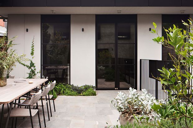A contemporary courtyard patio beside a modern residential facade, featuring a long outdoor dining table with woven chairs, pale stone paving, dark-framed glazed doors and windows, white rendered walls, black metal screening, and lush planted garden beds.
