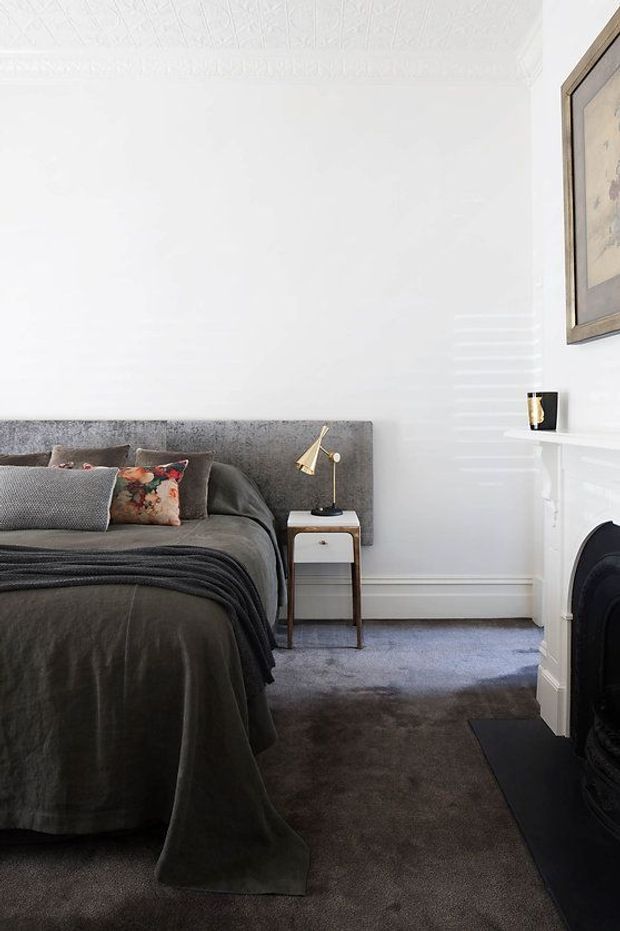 A refined bedroom interior with a grey upholstered bedhead, dark grey bedding and throw, patterned and textured cushions, and a small timber-and-white bedside table with a brass reading lamp. The room has white walls with ornate decorative ceiling detailing, a white fireplace mantel and dark fireplace surround, grey carpet, and framed artwork. The styling is calm, layered, and traditionally influenced with contemporary furnishings.