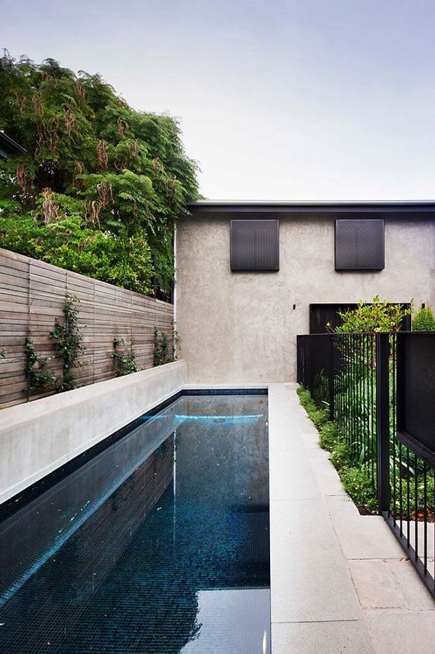 A narrow contemporary courtyard pool enclosed by rendered walls, horizontal timber screening, and black metal fencing. The rectangular pool has dark blue mosaic tiling and is bordered by pale concrete or stone paving. Dense greenery softens the boundary, while a simple flat-roofed rendered building with dark square window shutters forms the backdrop.