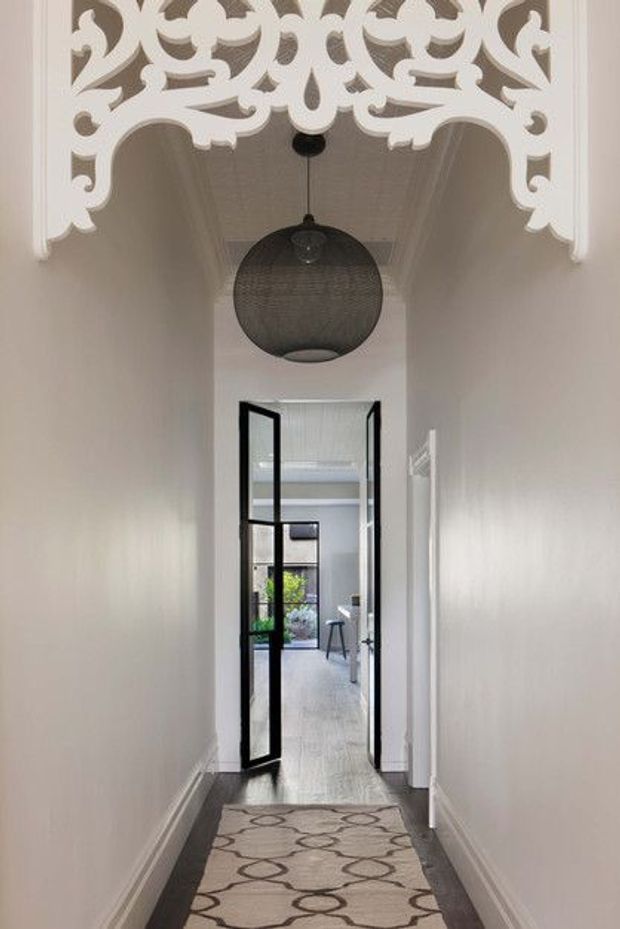 A narrow residential hallway with pale walls, dark timber flooring, and a patterned runner leading through black-framed glazed double doors into a brighter room. The hallway features ornate white decorative ceiling cornices and a large dark spherical pendant light, creating a refined transitional space with a contrast between traditional detailing and contemporary steel-framed doors.