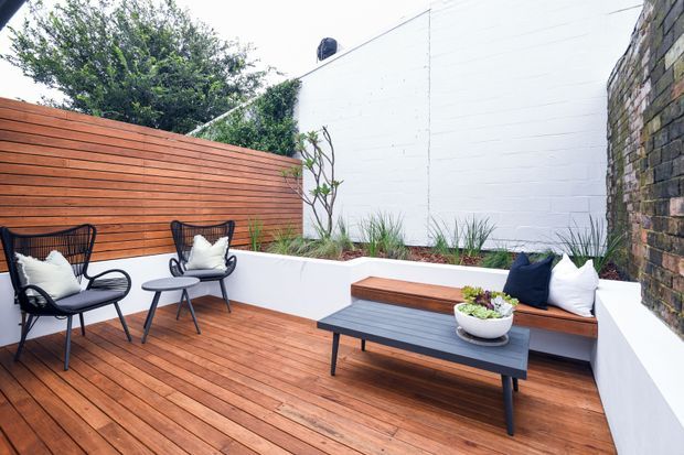 A compact landscaped outdoor courtyard terrace with warm timber decking, a horizontal timber privacy fence, white rendered planter and bench walls, and a dark brick boundary wall. Black outdoor lounge chairs and a low coffee table create a relaxed seating area, with integrated planting and built-in timber seating.