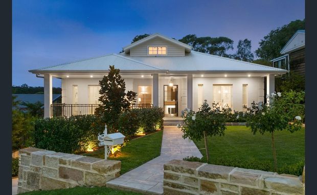 Front exterior view of a single-storey traditional house at dusk, featuring a broad hipped metal roof with a small upper gable dormer, deep covered verandah, pale weatherboard-style walls, illuminated windows, landscaped lawn and garden beds, and low stone boundary walls.
