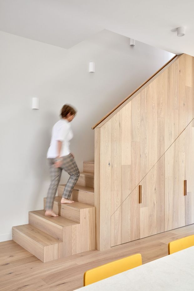Bright contemporary interior showing a timber staircase with integrated full-height storage cabinetry beneath and beside the stair. Pale oak-toned timber flooring, stair treads, wall panelling and cabinetry contrast with smooth white walls and ceiling. Minimal white wall and ceiling lights are visible, with yellow dining chairs and part of a light table in the foreground. A person is walking up the stairs.