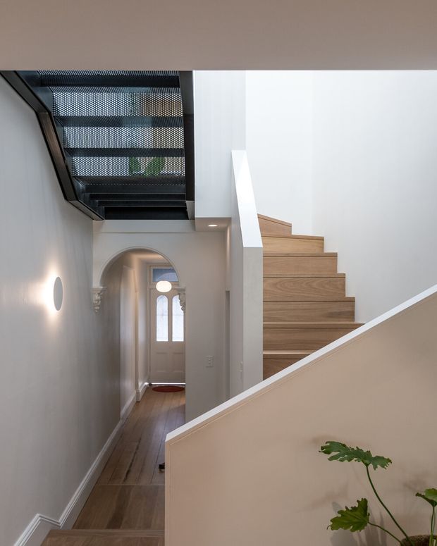 A bright, refined residential hallway and stairway with warm timber flooring and open-riser timber stairs. The space features white painted walls, classic architrave and cornice detailing, a decorative arched doorway at the end of the hall, and a dark perforated skylight or overhead screen bringing filtered light into the stairwell. A simple wall sconce and indoor greenery add subtle character.