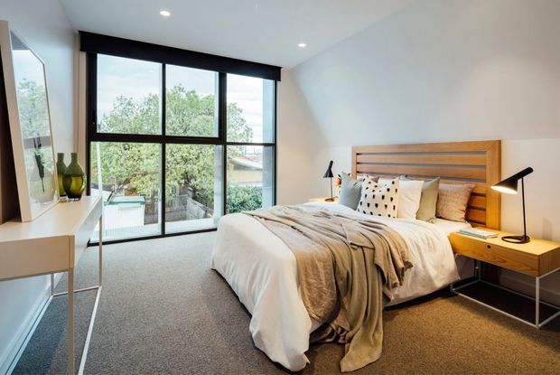 A bright contemporary bedroom with a large black-framed floor-to-ceiling window, warm timber slatted bedhead, bedside tables with lamps, light bedding, and soft grey carpet. A minimalist white dressing or storage unit sits along the left wall, with greenery visible outside.