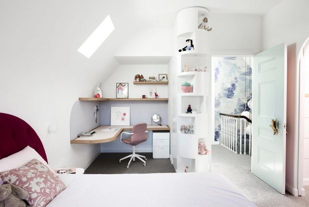 Bright children's bedroom with a built-in study nook, pale timber desk, shelving, swivel chair, rounded white display shelving, and an open door leading to a nursery or adjoining room. The room has white walls, carpeted flooring, soft furnishings, and a skylight providing strong natural light.