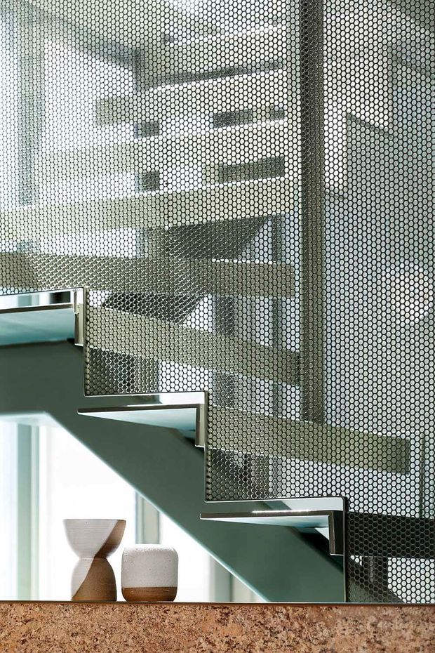 Close-up interior view of a contemporary staircase with floating timber treads, dark metal edging, and a perforated circular-pattern metal balustrade. The stair rises alongside a bright glazed area, with a stone or terrazzo-like ledge and decorative vessels in the foreground.