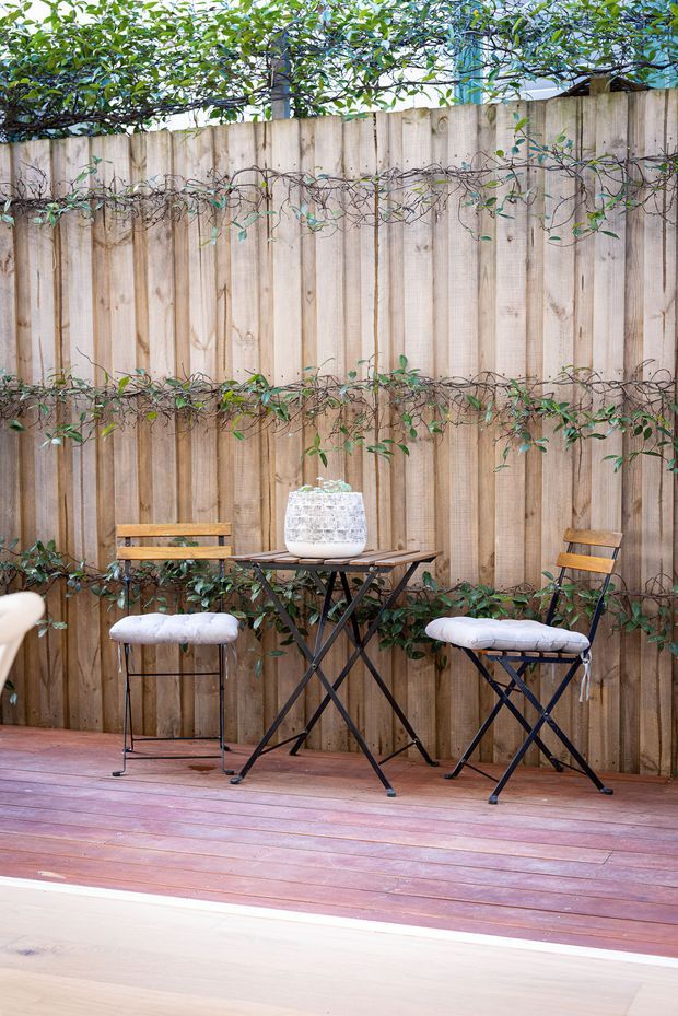 A compact outdoor entertaining area with a timber deck, a tall vertical timber privacy fence, and climbing vines trained along the fence. A small black folding table with a white textured planter sits between two black-framed stools or bistro chairs with wooden backrests and white cushions. The scene has a warm, natural garden-courtyard character.