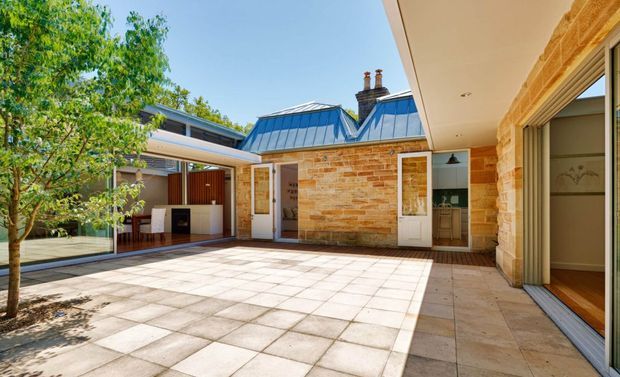 Sunlit residential courtyard framed by a brick house and covered outdoor living area. The space features pale square-paver flooring, warm exposed brick walls, blue-grey metal roof sections, timber detailing, a built-in outdoor kitchen or barbecue zone, and large glazed sliding doors opening into the home. Mature greenery borders the courtyard.