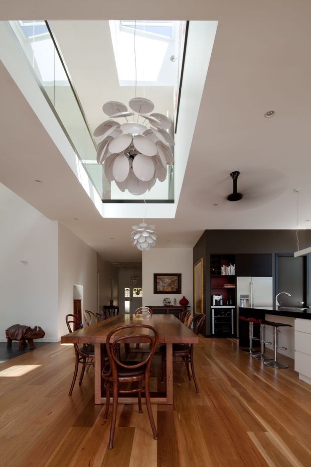 Bright open-plan dining and kitchen interior with a long timber dining table, curved timber dining chairs, dark built-in kitchen cabinetry, stainless-steel appliances, and timber flooring. A dramatic double-height skylight void with glass balustrade and sculptural pendant lighting forms the main architectural feature.