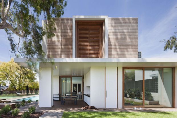 A contemporary two-storey residence viewed from the landscaped rear garden. The white lower level has broad roof overhangs, full-height timber-framed glazing, sliding doors and an outdoor dining area, while the upper level is clad in horizontal timber boards with a projecting framed opening containing timber louvres. A swimming pool and planted garden are visible to the side, with mature trees casting shadows across the facade.