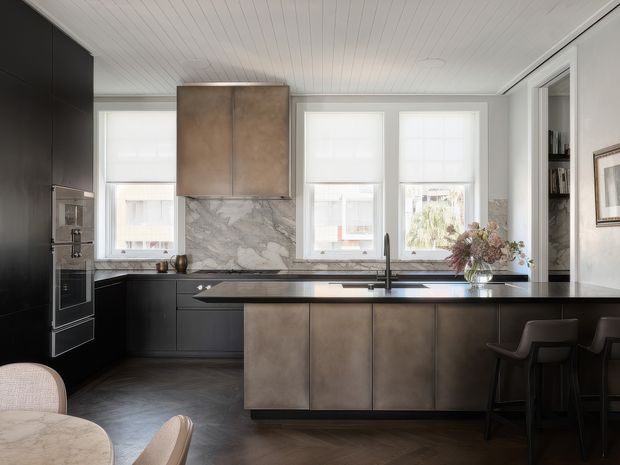 A sophisticated contemporary kitchen with dark matte cabinetry, a large central island, integrated appliances, and a sculptural metallic rangehood. The island and benchtops are dark and streamlined, while a veined stone backsplash adds texture behind the cooktop. Tall windows bring in abundant natural light, and a small adjoining dining area with bar stools is visible at the right. The ceiling has white painted timber boards, and the overall palette is dark, neutral, and refined.