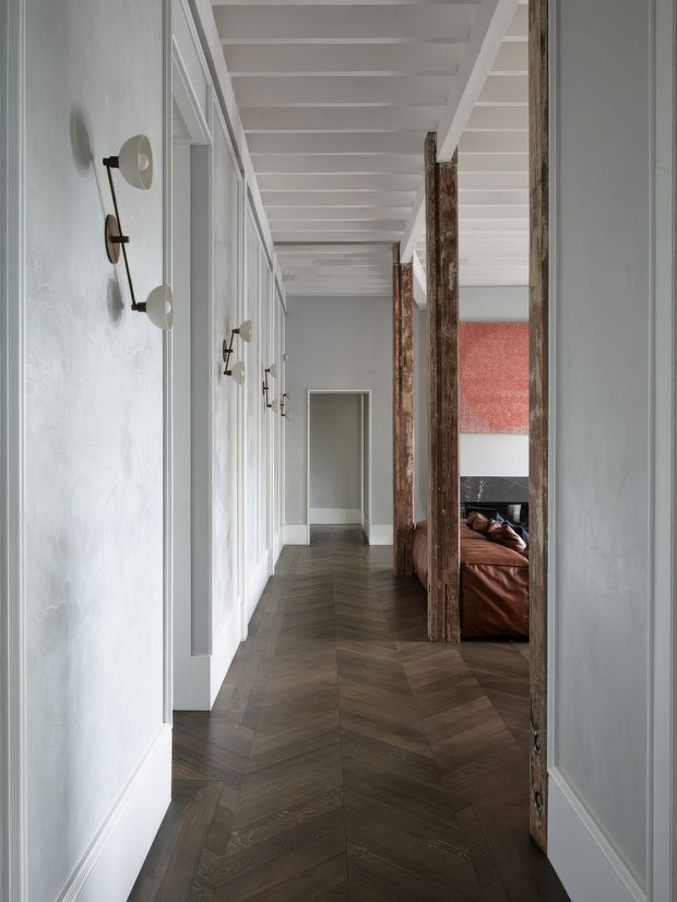 A long, refined interior hallway with dark herringbone timber flooring, pale grey paneled walls, white-painted exposed ceiling beams, and a series of wall-mounted sconces. Rustic timber posts and a partial view of a brown leather sofa and red artwork are visible through an opening on the right, creating a contrast between traditional detailing and contemporary furnishings.