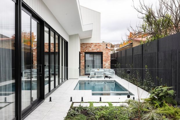 Contemporary residential courtyard viewed from an outdoor paved area, featuring a compact swimming pool, white rendered and red-brick building elements, extensive black-framed glazing, a dark vertical privacy fence, and landscaped planting. The space has a clean, refined modern aesthetic with a narrow urban courtyard layout.