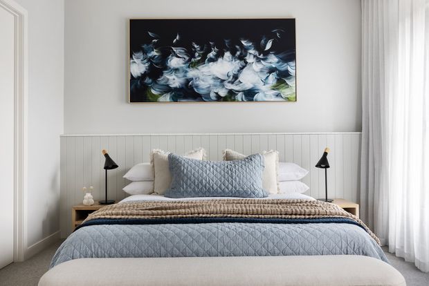 A bright, refined bedroom featuring a neatly layered bed with pale blue quilted bedding, textured neutral throws, and multiple pillows. The bed is set against a white vertical-panelled feature wall with slim black bedside lamps, while a blue-and-white abstract artwork provides a focal point above the headboard. Sheer white curtains soften the light at the window.