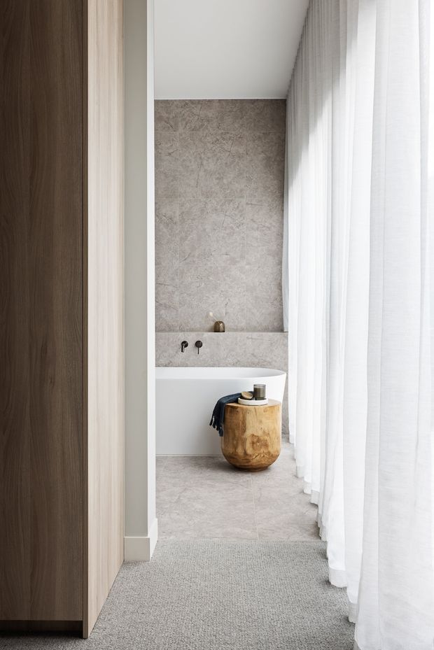 A serene, contemporary bathroom viewed from an adjoining carpeted space. The room features a freestanding white bathtub against a full-height mottled stone-look wall, pale stone-look flooring, dark minimal tapware, sheer white floor-to-ceiling curtains, and a rustic timber stool beside the bath. A dark timber door and light timber wall panel frame the opening.
