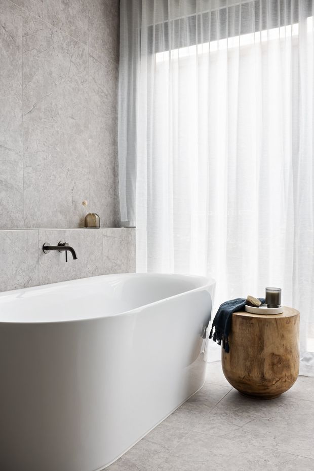 Bright, refined bathroom featuring a sculptural freestanding white bathtub, pale grey marble-look wall and floor tiles, a wall-mounted matte black bath tap, and sheer full-height white curtains beside a large window. A rounded natural timber stool with a dark textile and candle adds warmth and contrast.