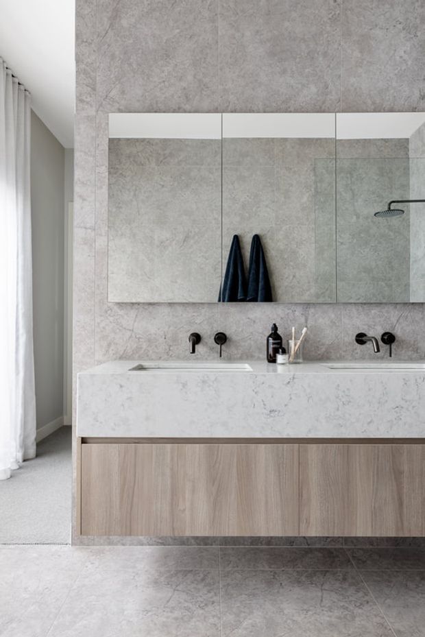 A refined contemporary bathroom featuring a floating double vanity with pale wood cabinetry, a thick light stone benchtop, wall-mounted black tapware, and a large mirrored cabinet. Grey stone-look wall and floor tiles create a cohesive monochromatic finish, with a walk-in shower visible at right and sheer curtains at left.
