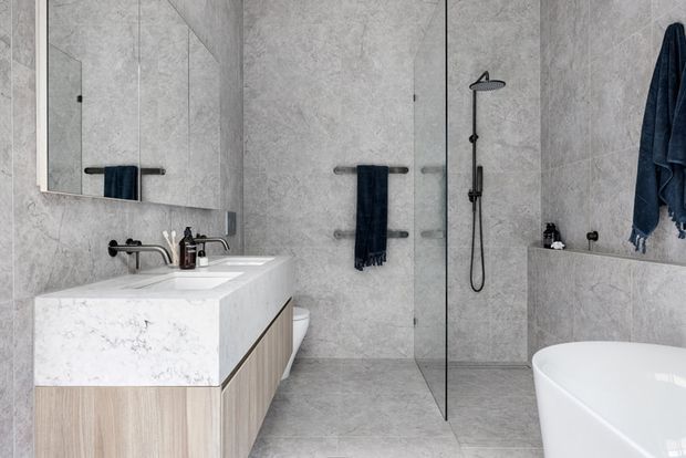 A contemporary bathroom in a restrained grey and white palette, featuring large-format grey stone-look wall and floor tiles, a floating light timber vanity with a white stone benchtop and integrated basin, black tapware and towel rails, a frameless glass shower, and a freestanding white bathtub. A mirrored wall cabinet reflects the vanity and shower area.