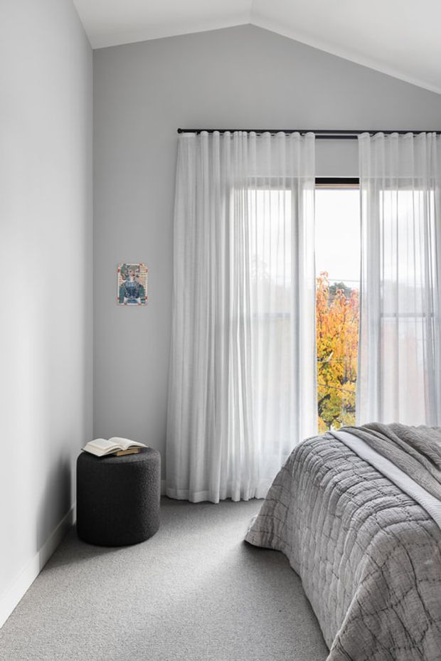 A quiet, contemporary bedroom with soft grey walls, a sloped ceiling, full-height sheer white curtains, and a large window overlooking autumn foliage. The room has light grey carpet, a partially visible bed dressed in a textured grey quilt, and a small dark upholstered cylindrical stool with an open book.