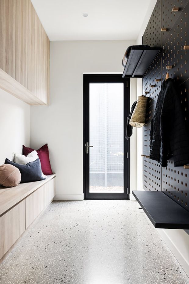 Bright, compact entryway with a built-in light timber bench and overhead cabinetry on the left, a dark perforated storage wall with hooks, shelf and bench on the right, and a black-framed glazed exterior door at the end. The floor is a light terrazzo-like finish, creating a clean, functional, contemporary mudroom-style space.