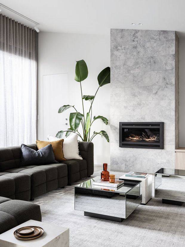 A bright, contemporary living room with a dark modular sectional sofa, mirrored rectangular coffee tables, a large grey area rug, sheer floor-to-ceiling curtains, indoor tropical plants, and a recessed fireplace set into a full-height grey stone-tile feature wall.