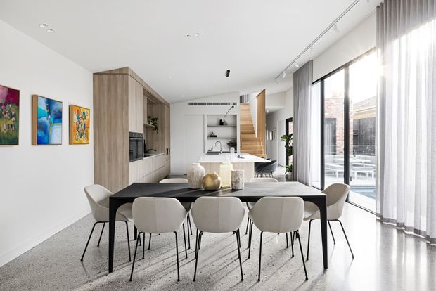 Bright open-plan contemporary interior with a dining area in the foreground and a compact kitchen behind it. The room features a long dark dining table with upholstered chairs, light timber built-in cabinetry, a white island and work surfaces, polished light terrazzo-like flooring, white walls, large floor-to-ceiling windows with sheer curtains, and colourful framed artwork.