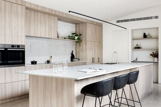 Bright contemporary kitchen featuring extensive light timber-grain cabinetry, a large pale stone island with integrated sink and seating for four, built-in black appliances, open shelving, and a minimalist linear pendant light. The space has a clean, refined, warm-neutral palette with subtle greenery and black accents.
