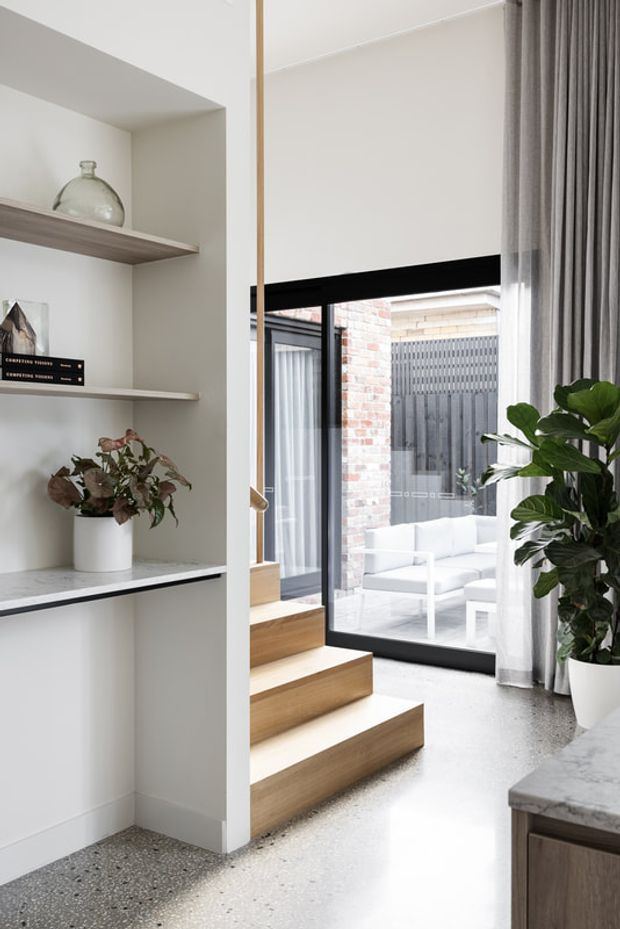Bright contemporary residential interior showing a compact stairway leading to a glazed courtyard or outdoor sitting area. Built-in white shelving with timber shelves and decorative objects occupies the left side, while black-framed floor-to-ceiling glazing, sheer curtains, terrazzo flooring, timber stair treads, and a large indoor plant define the space. A stone-topped kitchen or console surface is visible in the foreground.